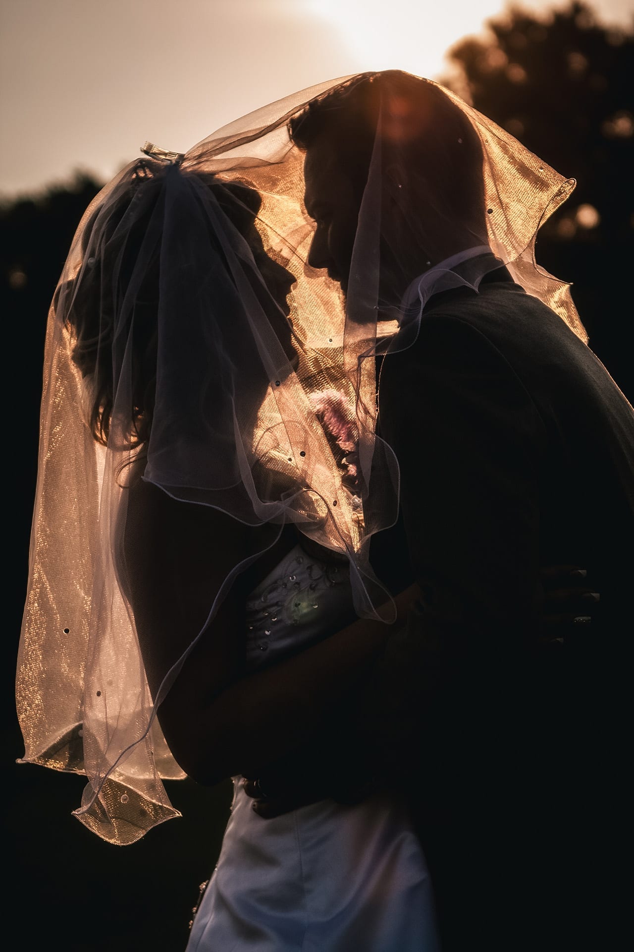 A newly married couple silhouetted together beneath a backlit wedding veil