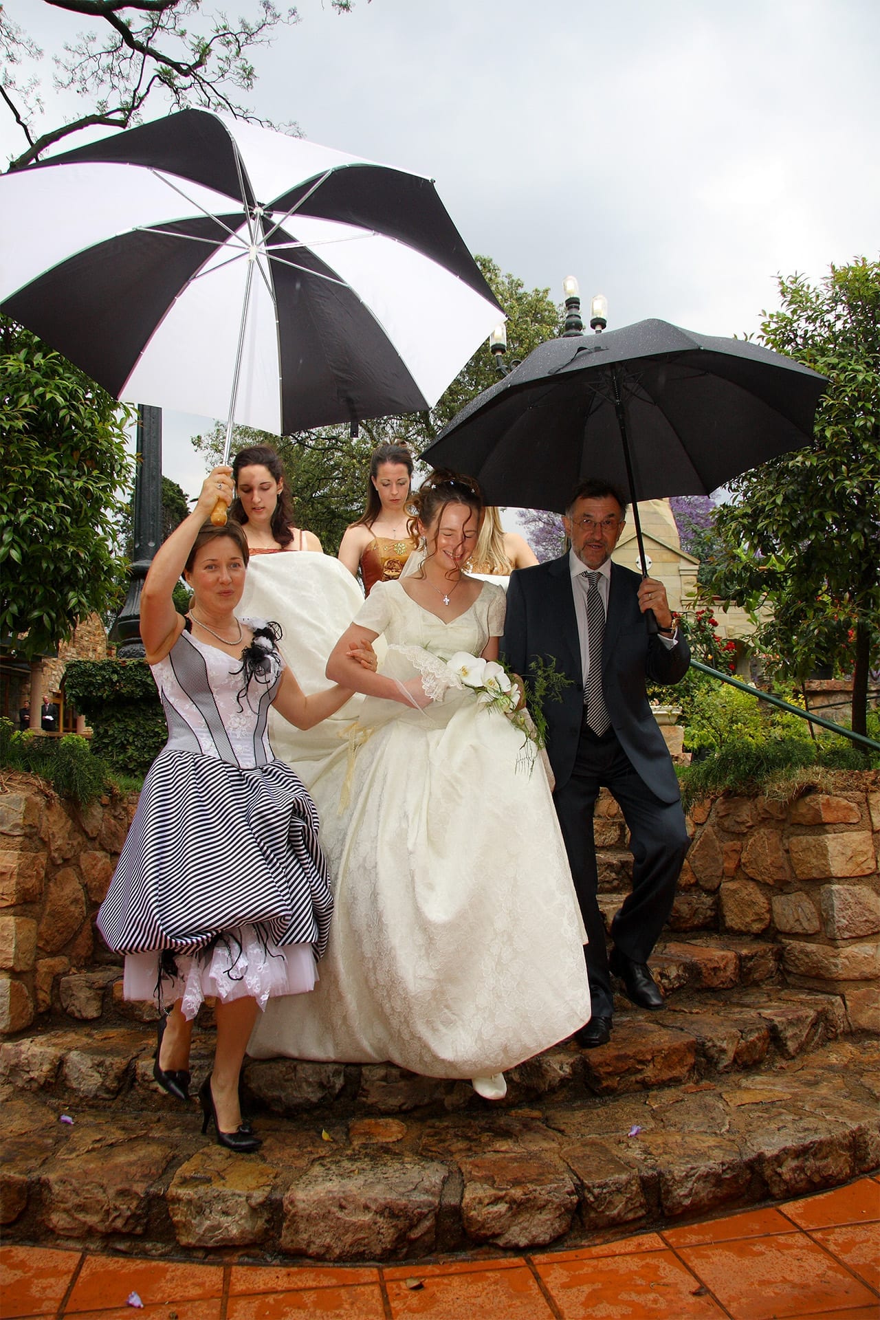 A bride walking carefully down wet stone steps while family shield her from the rain with umbrellas